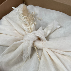 Aerial photo of the inside of a cardboard box. Contents wrapped in natural muslin fabric tied using the furoshiki method in the centre of the box. A small bunch of natural dried flowers appear placed within the tied fabric in the centre.